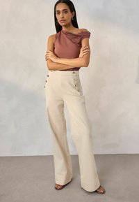 Bruine top met één schouder en draping, gecombineerd met hooggesneden, crèmekleurige, wijde broek met knoopdetails aan de zijkant.
