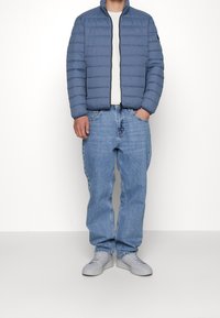 Giacca puffy azzurra con cuciture orizzontali, chiusura con zip e colletto alto, abbinata a jeans azzurri e sneakers grigie.