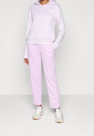 Persona vistiendo un chándal Adidas color morado claro con sudadera con capucha y cremallera, y pantalones a juego, combinado con zapatillas blancas Adidas.
