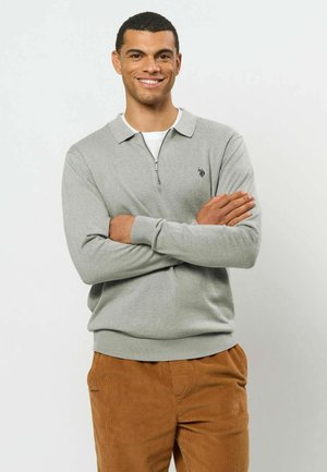 Homme souriant, bras croisés, portant un polo gris clair à manches longues et un pantalon en velours côtelé marron sur un fond uni.