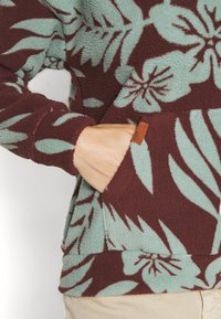 Main dans la poche d'un pull en polaire avec un motif floral bordeaux et bleu clair, montrant un tissu texturé et une petite étiquette marron de la marque.