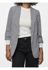Blazer de patrón de houndstooth en blanco y negro con cuello chal, mangas enrolladas y abertura frontal, combinado con una blusa clara y pantalones negros.