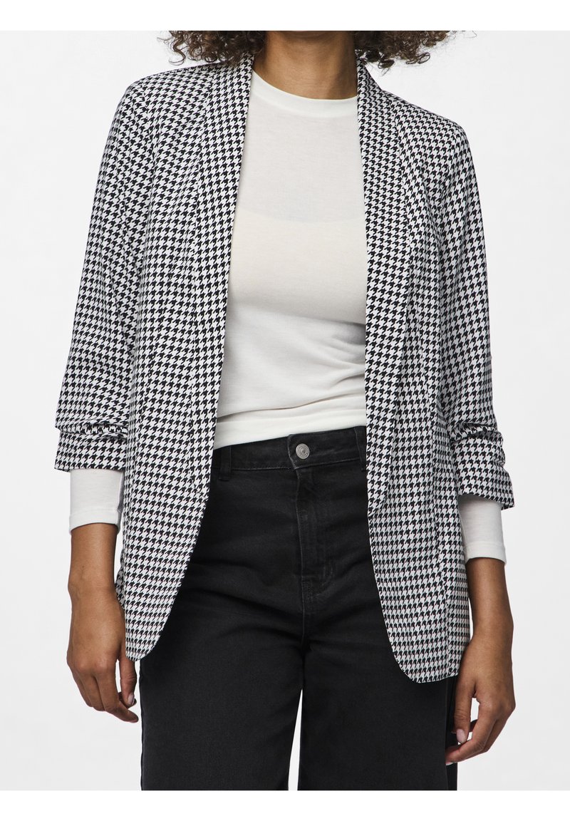 Blazer de patrón de houndstooth en blanco y negro con cuello chal, mangas enrolladas y abertura frontal, combinado con una blusa clara y pantalones negros.