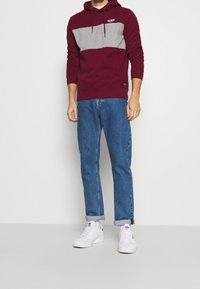 Sudadera con capucha en color burdeos y gris con un diseño de bloques de color, combinada con jeans rectos azules y zapatillas blancas. Estilo casual y cómodo.