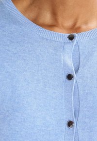 Ljusblå cardigan med ribbad halsringning, prydd med tre svarta knappar fram. Tyget ser mjukt och texturerat ut.