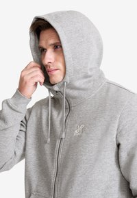 Grå zip-up hoodie med hög krage och justerbara dragsnören. Har en liten logotyp på bröstet och ett mjukt, texturerat tyg.