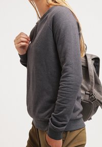Felpa grigia in un morbido misto cotone con scollatura a girocollo, polsini a coste e un dettaglio del logo minimale sul petto. Abbinata a pantaloni beige.
