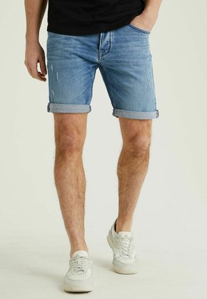 Homme portant un short en denim bleu retroussé, des baskets blanches et une chemise noire, debout avec une main dans la poche devant un fond uni.