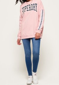 Sudadera de color rosa claro con "SUPERDRY" en azul marino, con mangas largas y detalle blanco en los laterales. Combinada con jeans ajustados de mezclilla azul y zapatillas blancas.