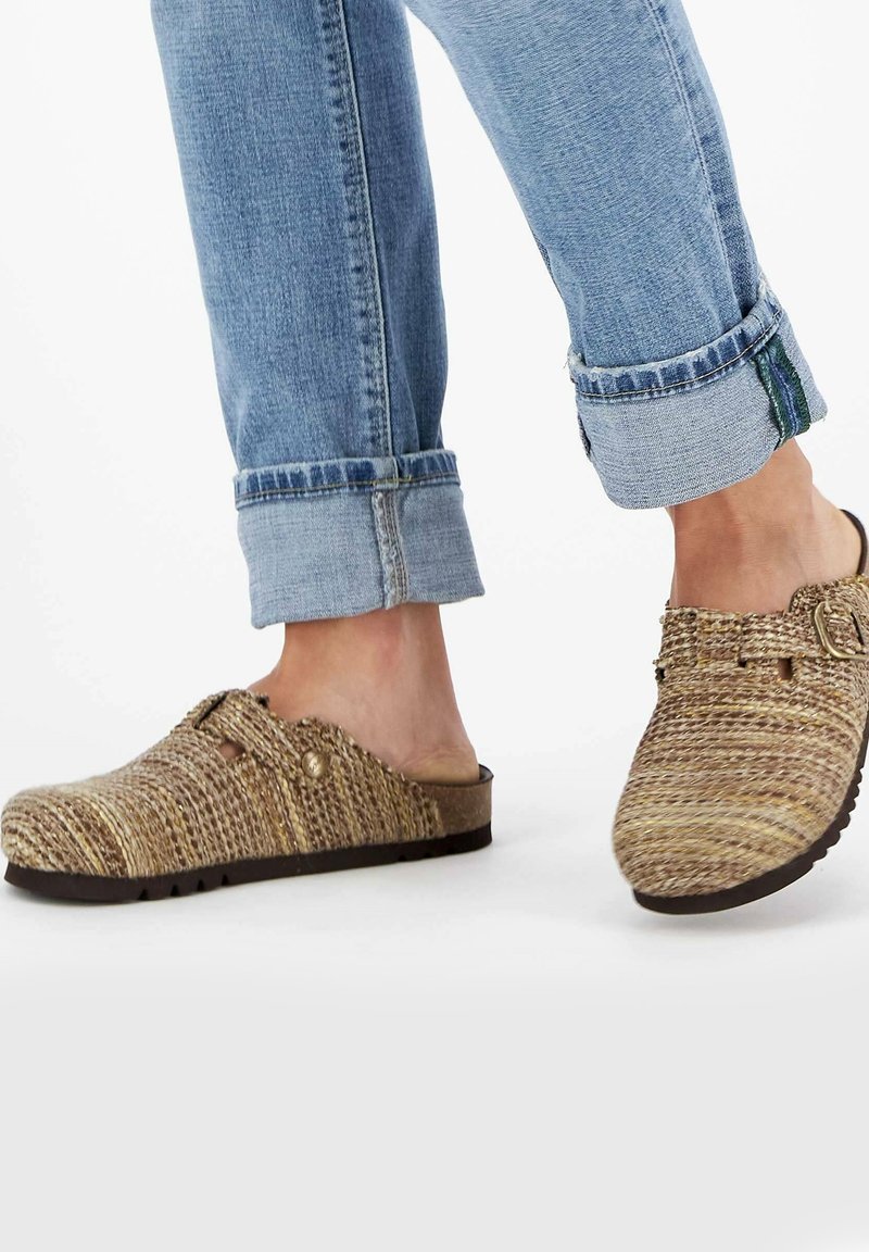 Sabots tissés, de couleur beige, avec un bout arrondi et une boucle ajustable, présentant une surface texturée et une semelle en caoutchouc marron. Pantalons en denim à revers visibles.