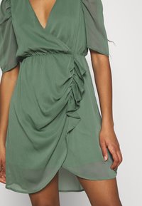 Robe enveloppante verte en tissu léger, avec un profond décolleté en V, des manches bouffantes et un design froncé sur le devant, présentant une finition légèrement transparente.