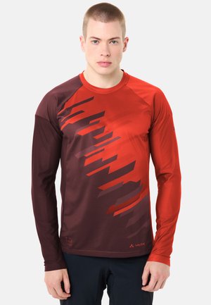 Jeune homme portant un maillot de sport à manches longues avec des motifs géométriques rouges et bordeaux foncé, debout devant un fond blanc uni.