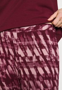 Burgundska majica u kombinaciji s hlačama s uzorkom i teksturom koje sadrže nijanse ružičaste i maroon, prikazujući kutne geometrijske dizajne.