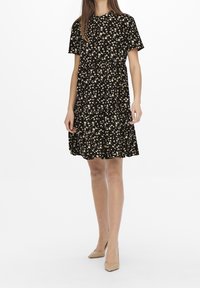 Abito nero a maniche corte con motivi floreali bianchi, gonna arricciata e scollatura rotonda. Indossato con tacchi beige. Tessuto dalla trama liscia.