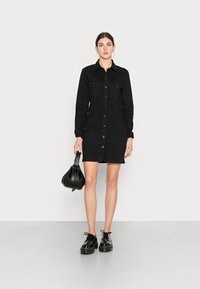 Vestido camiseiro de denim preto com mangas longas, frente abotoada, dois bolsos no peito e corte reto. Sapatos plataforma pretos e bolsa.