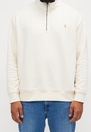 Mężczyzna w jasnobeżowym pulowerze z quarter-zip z żółtym logo Polo na lewej piersi i niebieskich dżinsach, stojący na neutralnym tle.