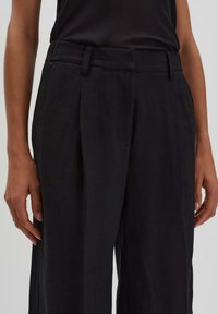 Black high-waisted trousers made of a textured fabric, featuring pleats, side pockets, and belt loops, with a straight-leg design.