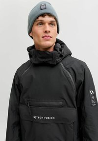 Noor mees kannab halli mütsi ja musta Tech Fusion jope, millel on ees lukuga tasku, seistes ühtlase heleda tausta eest.