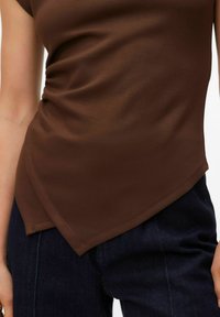 Haut marron sans manches en tissu lisse, avec un ourlet asymétrique et un léger froncé sur un côté. Porté avec un pantalon en denim foncé.