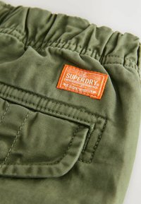 Zelene pamučne kratke hlače s elastičnom waistbands, s jednom džepom i narančastom oznakom "SUPERDRY" ušivenom na bočnoj strani.