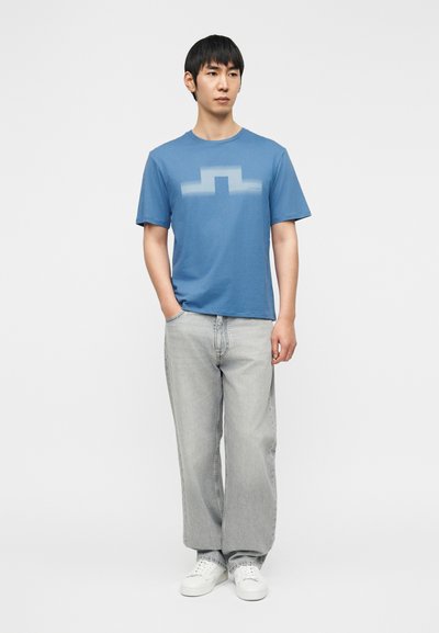 Μπλε κοντομάνικο μπλουζάκι με σχέδιο σε κλίση στο στήθος, συνδυασμένο με ανοιχτό γκρι τζιν παντελόνι φαρδιάς γραμμής και λευκά αθλητικά παπούτσια.