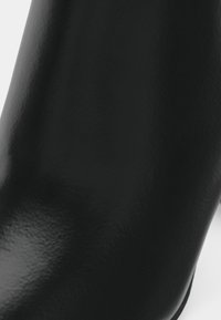 Detailní záběr na hladký, lesklý černý kožený povrch s jemnou strukturou a měkkým zakřiveným tvarem na světlém pozadí.