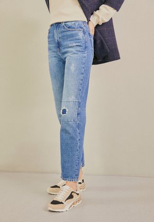 Osoba nosząca jasnoniebieskie podarte dżinsy z łatą na kolanie oraz beżowo-czarne sneakersy, z ręką w kieszeni, na jednolitym beżowym tle.