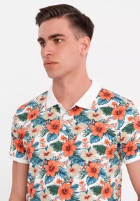 Blommig polo skjorta med livfulla orange hibiskusar och gröna löv på en vit bakgrund, med en kontrasterande vit krage och tre knappar.