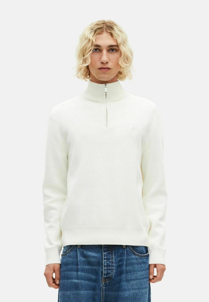 Weißer Strickpullover mit hohem Kragen und halbem Reißverschluss. Verfügt über gerippte Bündchen und Saum, kombiniert mit blauer Jeans. Einfaches, klares Design.