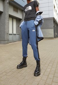 Svart T-shirt med vitt logotyp, blå joggers med svarta sidor, och svarta kängor. Strukturerad tyg, avslappnad passform.