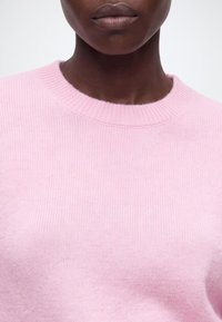 Gros plan d'une personne portant un pull tricoté rose clair à encolure ronde, montrant la partie inférieure du visage et le cou.
