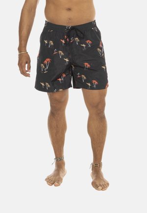 Homme portant un short de bain noir orné d'un motif tropical de planche de surf et de palmiers, debout pieds nus avec des bracelets de cheville aux deux jambes.