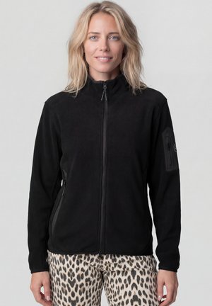 Frau mit blonden Haaren, die eine schwarze Fleecejacke mit Reißverschluss und Leopardenmuster-Hose trägt, steht vor einem schlichten hellen Hintergrund.