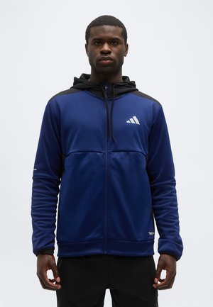 Muž v modro-čiernej mikine s kapucňou a so zipsom stojí pred jednoduchým svetlým pozadím.