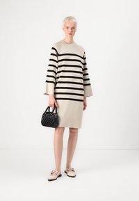 Beige, stripete strikkekjole med svarte striper, lange ermer og rund hals. Kombinert med en liten svart strukturert veske og lyse sko.