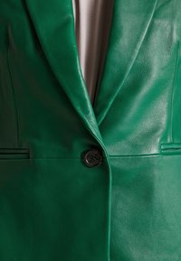 Blazer en cuir vert avec un seul bouton noir fermé et un tissu beige visible sous les revers.