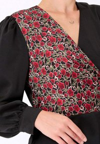 Robe noire avec un tissu à motifs floraux rouges sur un côté du décolleté en V profond et des manches bouffantes ; des points délicats mettent en valeur le design.