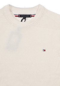 Pullover aus gestricktem Material in Cremefarbe mit Rundhalsausschnitt, verziert mit einem kleinen gestickten Logo in Rot, Weiß und Blau auf der Brust.