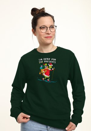 Groen sweatshirt met een kleurrijke illustratie van de Grinch die skiet met cadeaus en de tekst "IK BEN HIER VOOR DE CADEAUS." Zachte stof, losse pasvorm.