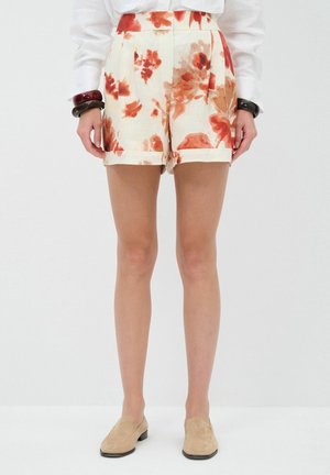 Ben iført cremefarvede shorts med rødt og orange blomstermønster, beige loafers, hvid langærmet skjorte og store mørke armringer på håndleddene.