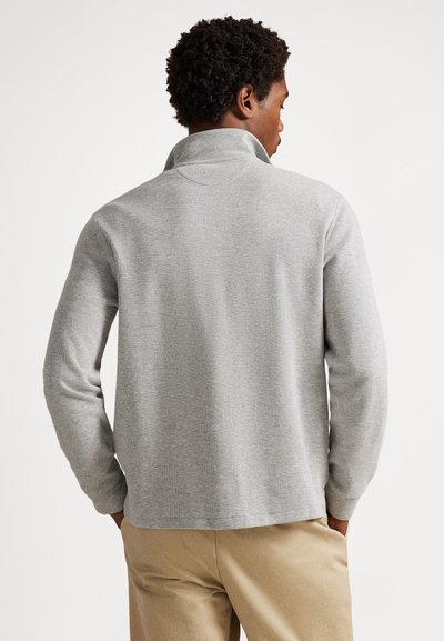 Svetlo sivi pulover z visokim ovratnikom, teksturirano tkanino in dolgimi rokavi. Ima sproščen kroj in ravno spodnjo rob.