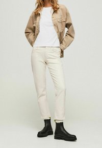 Beige jacka över en vit skjorta, kombinerad med krämb färgade jeans och svarta chelsea-boots med texturerade sulor. Enkel och funktionell design.