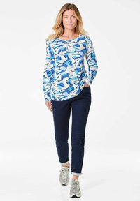 Langärmliges Oberteil in einem geometrischen Muster aus Blau, Weiß und Hellblau; kombiniert mit dunkelblauen Slim-Fit-Jeans und silbernen Sneakers.