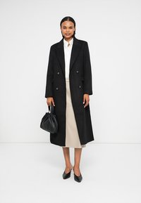 Sort uld dobbeltbrystet frakke med skræddersyet pasform, båret over en beige nederdel. Sort håndtaske og sko fuldender looket. Ensartet baggrund.