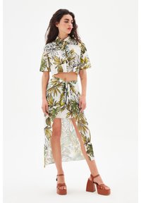 Witte crop top en rok set met bloemenprint, featuring groene en gele bladeren, strik taille en doorzichtig materiaal. Inclusief bruine plateau schoenen.