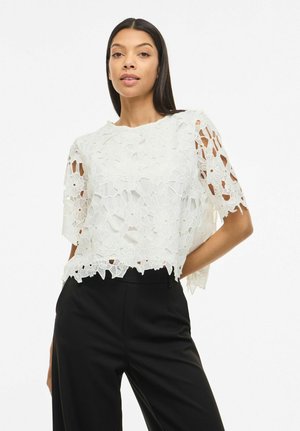 Kobieta o długich ciemnych włosach nosi biały, kwiecisty koronkowy top z krótkim rękawem i czarne spodnie z wysokim stanem, pozując z jedną ręką na biodrze.