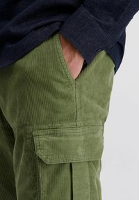 Hand in de zak van olijfgroene corduroy cargo broek, gedragen met een donkerblauw denim shirt.