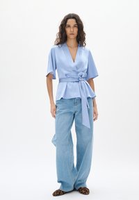 Blusa avvolgente azzurro chiaro con maniche corte e un fiocco in vita, abbinata a jeans denim larghi e mules con stampa leopardata.