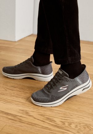 Graue Sportschuhe mit einem Obermaterial aus Mesh, strukturiertem Design, runder Zehenpartie und gepolstertem weißen Absatz. Sie verfügen über Schnürsenkel und subtile Markierungsakzente.