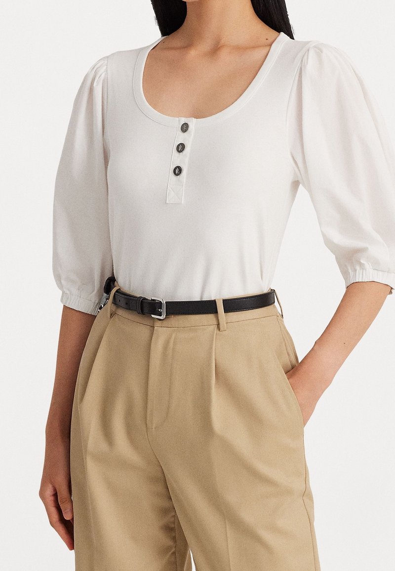 White short-sleeve top with a scooped neckline, three black buttons, and gathered puff sleeves, paired with high-waisted beige trousers and a black belt.
